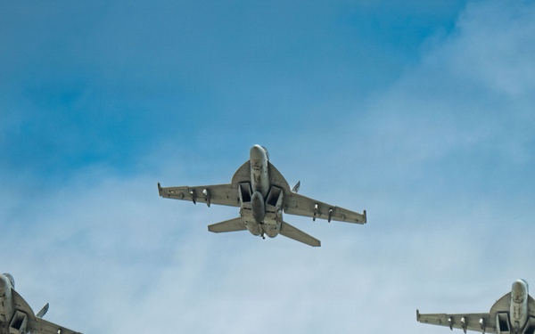 Three military jet aircraft are flying in a triangular formation against a partly cloudy blue sky. The aircraft are viewed from below, revealing their undersides, wing configurations, and hardpoints with mounted equipment. The formation appears to be a standard echelon or delta flight pattern consistent with military aviation operations.