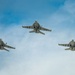 Three military jet aircraft are flying in a triangular formation against a partly cloudy blue sky. The aircraft are viewed from below, revealing their undersides, wing configurations, and hardpoints with mounted equipment. The formation appears to be a standard echelon or delta flight pattern consistent with military aviation operations.