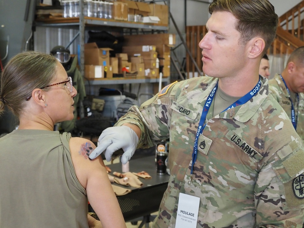 A U.S. Army soldier identified by name tape as 'CARO' with the rank of Staff Sergeant is applying moulage (simulated wound makeup) to a female soldier's upper arm during a training exercise. The soldier is wearing latex gloves and has a lanyard badge reading 'MOULAGE,' indicating his role in the exercise. The setting appears to be a warehouse or storage facility used as a training area.