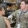 A U.S. Army soldier identified by name tape as 'CARO' with the rank of Staff Sergeant is applying moulage (simulated wound makeup) to a female soldier's upper arm during a training exercise. The soldier is wearing latex gloves and has a lanyard badge reading 'MOULAGE,' indicating his role in the exercise. The setting appears to be a warehouse or storage facility used as a training area.