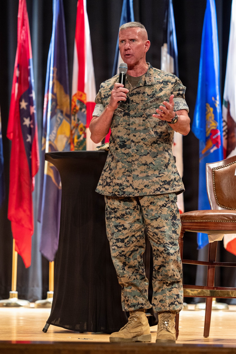 A U.S. Marines officer in MARPAT digital camouflage uniform is speaking on a stage while holding a handheld microphone. He appears to be addressing an audience at a formal military event or conference, gesturing with his free hand. Multiple U.S. state and military flags are displayed on stands behind him.