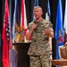 A U.S. Marines officer in MARPAT digital camouflage uniform is speaking on a stage while holding a handheld microphone. He appears to be addressing an audience at a formal military event or conference, gesturing with his free hand. Multiple U.S. state and military flags are displayed on stands behind him.