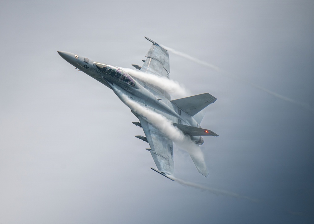 A twin-engine fighter jet, identifiable as an F/A-18 Super Hornet, is captured in a high-speed, banked maneuver against an overcast gray sky. Dramatic vapor condensation trails (Prandtl-Glauert condensation) are visibly streaming across the fuselage, wings, and leading edges due to rapid pressure changes at high speed. The aircraft bears the number '104' on its nose and appears to be in a steep climbing or rolling attitude.