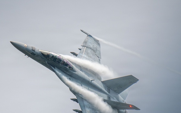 A twin-engine fighter jet, identifiable as an F/A-18 Super Hornet, is captured in a high-speed, banked maneuver against an overcast gray sky. Dramatic vapor condensation trails (Prandtl-Glauert condensation) are visibly streaming across the fuselage, wings, and leading edges due to rapid pressure changes at high speed. The aircraft bears the number '104' on its nose and appears to be in a steep climbing or rolling attitude.