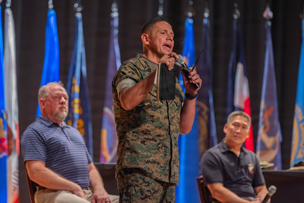 A U.S. Marine in woodland MARPAT camouflage uniform is standing and speaking into a microphone at what appears to be a formal military briefing or town hall event. Two civilians in casual attire are seated behind him on a stage. Multiple military and organizational flags are displayed in the background.