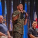 A U.S. Marine in woodland MARPAT camouflage uniform is standing and speaking into a microphone at what appears to be a formal military briefing or town hall event. Two civilians in casual attire are seated behind him on a stage. Multiple military and organizational flags are displayed in the background.