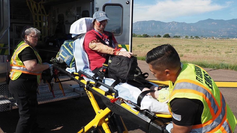 Two emergency medical personnel in high-visibility yellow vests are attending to a patient on a yellow Stryker stretcher near the rear of an ambulance. The patient, wearing a red shirt and blue cap, appears to have simulated injuries (moulage makeup) on their face and arm, suggesting this is a training exercise or drill. The scene is set outdoors in what appears to be a flat, open area with mountains visible in the background.
