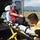 Two emergency medical personnel in high-visibility yellow vests are attending to a patient on a yellow Stryker stretcher near the rear of an ambulance. The patient, wearing a red shirt and blue cap, appears to have simulated injuries (moulage makeup) on their face and arm, suggesting this is a training exercise or drill. The scene is set outdoors in what appears to be a flat, open area with mountains visible in the background.