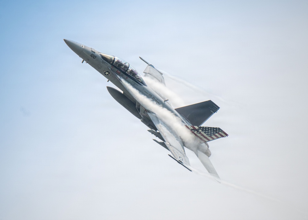 A U.S. Navy F/A-18 Super Hornet fighter jet is captured in a steep climbing maneuver against a pale blue sky. The aircraft bears the number '100' on its fuselage and features a distinctive American flag livery on its wing. Visible vapor condensation trails stream across the fuselage, indicating high-speed, high-G flight conditions.