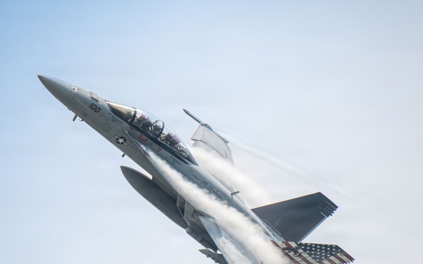 A U.S. Navy F/A-18 Super Hornet fighter jet is captured in a steep climbing maneuver against a pale blue sky. The aircraft bears the number '100' on its fuselage and features a distinctive American flag livery on its wing. Visible vapor condensation trails stream across the fuselage, indicating high-speed, high-G flight conditions.