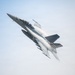 A U.S. Navy F/A-18 Super Hornet fighter jet is captured in a steep climbing maneuver against a pale blue sky. The aircraft bears the number '100' on its fuselage and features a distinctive American flag livery on its wing. Visible vapor condensation trails stream across the fuselage, indicating high-speed, high-G flight conditions.