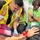 This image depicts a medical training or emergency drill scenario taking place inside an ambulance. A person wearing a high-visibility vest and using a stethoscope is attending to a simulated patient who has theatrical makeup applied to simulate injuries such as burns and wounds. Several observers wearing green shirts and lanyards with 'FLY COS' badges are present, suggesting this is an organized training exercise or public demonstration.