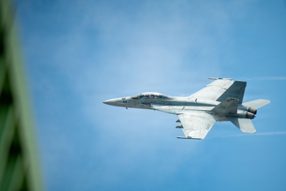 A military jet fighter, marked with the number '104', is captured in flight against a clear blue sky. The aircraft appears to be a Boeing F/A-18 Hornet or Super Hornet based on its twin-engine configuration and distinctive twin-tail design. The photo is taken from a slightly elevated angle, possibly from another aircraft or elevated vantage point, with a blurred green element visible in the foreground left.