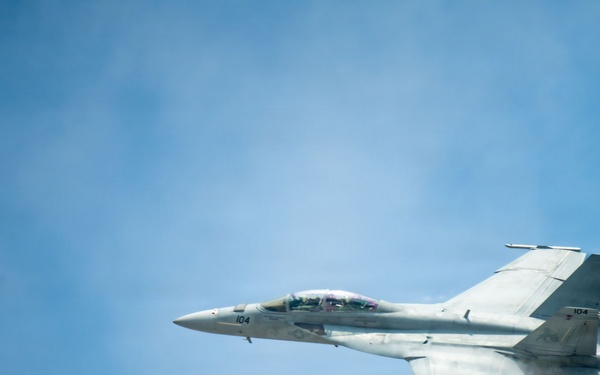 A military jet fighter, marked with the number '104', is captured in flight against a clear blue sky. The aircraft appears to be a Boeing F/A-18 Hornet or Super Hornet based on its twin-engine configuration and distinctive twin-tail design. The photo is taken from a slightly elevated angle, possibly from another aircraft or elevated vantage point, with a blurred green element visible in the foreground left.