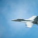 A military jet fighter, marked with the number '104', is captured in flight against a clear blue sky. The aircraft appears to be a Boeing F/A-18 Hornet or Super Hornet based on its twin-engine configuration and distinctive twin-tail design. The photo is taken from a slightly elevated angle, possibly from another aircraft or elevated vantage point, with a blurred green element visible in the foreground left.