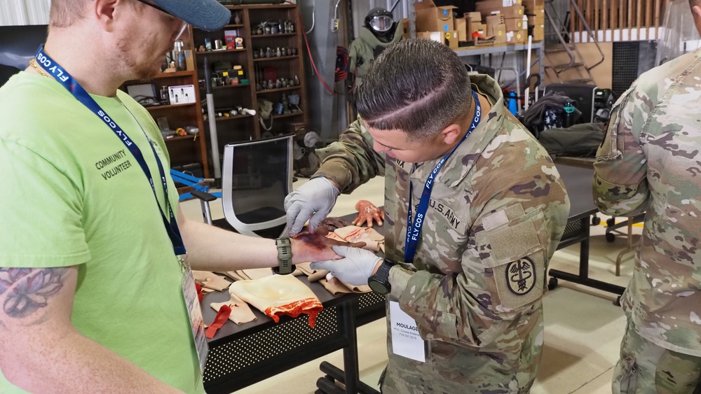 A U.S. Army soldier wearing camouflage uniform and latex gloves is applying moulage (simulated wound makeup) to a community volunteer's arm during what appears to be a training exercise. The soldier's badge reads 'MOULAGE' and both individuals are wearing blue 'FLYCOS' lanyards. The setting appears to be an indoor training facility or warehouse with shelving and equipment in the background.