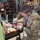 A U.S. Army soldier wearing camouflage uniform and latex gloves is applying moulage (simulated wound makeup) to a community volunteer's arm during what appears to be a training exercise. The soldier's badge reads 'MOULAGE' and both individuals are wearing blue 'FLYCOS' lanyards. The setting appears to be an indoor training facility or warehouse with shelving and equipment in the background.