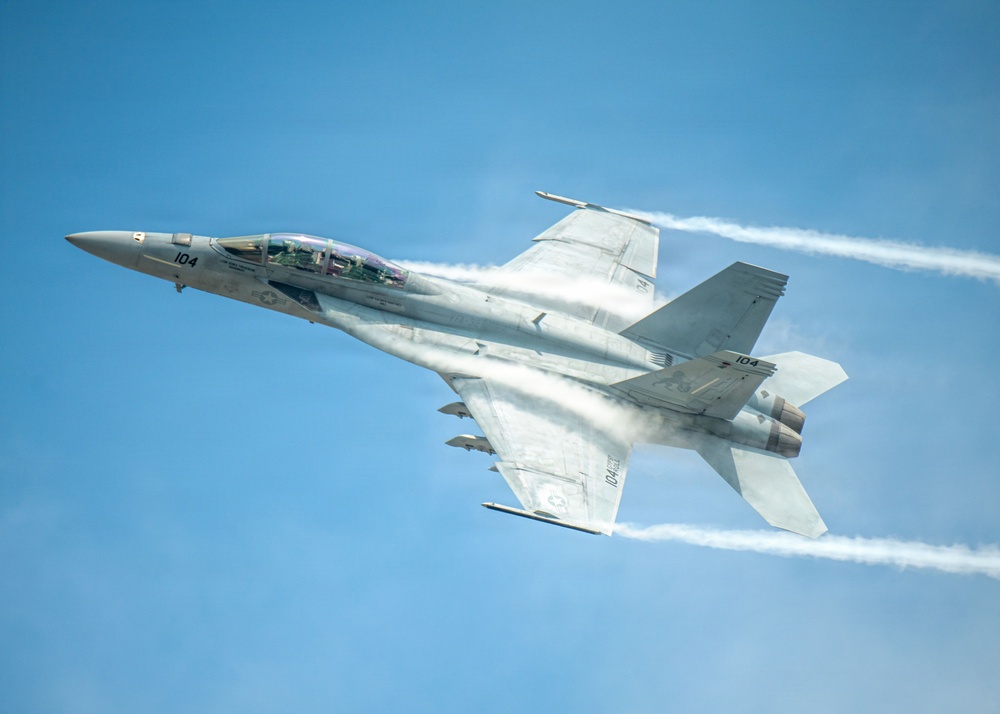 A U.S. Navy F/A-18F Super Hornet fighter jet, marked with the number '104', is captured in flight against a clear blue sky. The aircraft is producing visible vapor condensation clouds around its fuselage and twin white contrails from its engines, indicating high-speed, low-altitude flight. The image is a high-quality aerial photograph likely taken at an airshow or during a demonstration flight.