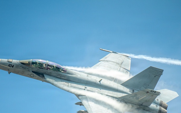 A U.S. Navy F/A-18F Super Hornet fighter jet, marked with the number '104', is captured in flight against a clear blue sky. The aircraft is producing visible vapor condensation clouds around its fuselage and twin white contrails from its engines, indicating high-speed, low-altitude flight. The image is a high-quality aerial photograph likely taken at an airshow or during a demonstration flight.