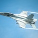 A U.S. Navy F/A-18F Super Hornet fighter jet, marked with the number '104', is captured in flight against a clear blue sky. The aircraft is producing visible vapor condensation clouds around its fuselage and twin white contrails from its engines, indicating high-speed, low-altitude flight. The image is a high-quality aerial photograph likely taken at an airshow or during a demonstration flight.
