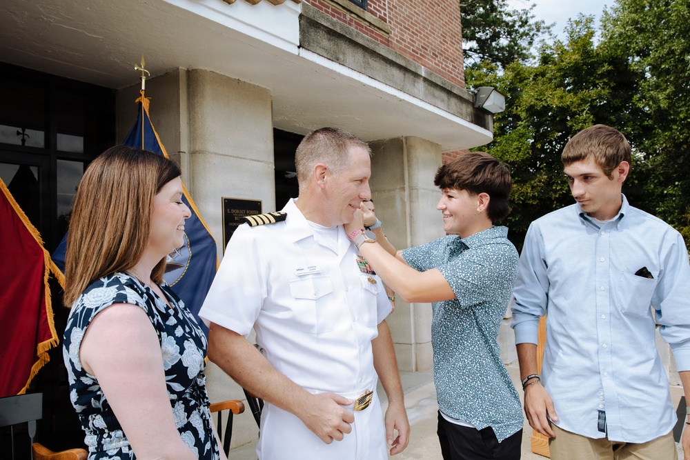 A U.S. Navy officer in white dress uniform is being pinned with insignia or medals by a younger person, likely a family member, during what appears to be a promotion or award ceremony. Three civilians — a woman in a floral dress and two younger males — are present, suggesting this is a family-attended military ceremony. The setting is outdoors in front of a brick building with military flags visible in the background.