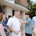 A U.S. Navy officer in white dress uniform is being pinned with insignia or medals by a younger person, likely a family member, during what appears to be a promotion or award ceremony. Three civilians — a woman in a floral dress and two younger males — are present, suggesting this is a family-attended military ceremony. The setting is outdoors in front of a brick building with military flags visible in the background.