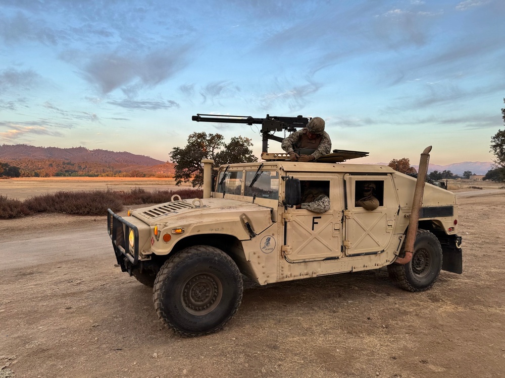A tan/desert-colored military HMMWV (Humvee) is parked on a dirt road or open terrain during early morning or evening light. At least two personnel in military uniforms and helmets are visible, one manning a crew-served weapon mounted on the roof turret. The setting appears to be a training environment in a semi-arid landscape with mountains and trees in the background.