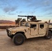 A tan/desert-colored military HMMWV (Humvee) is parked on a dirt road or open terrain during early morning or evening light. At least two personnel in military uniforms and helmets are visible, one manning a crew-served weapon mounted on the roof turret. The setting appears to be a training environment in a semi-arid landscape with mountains and trees in the background.