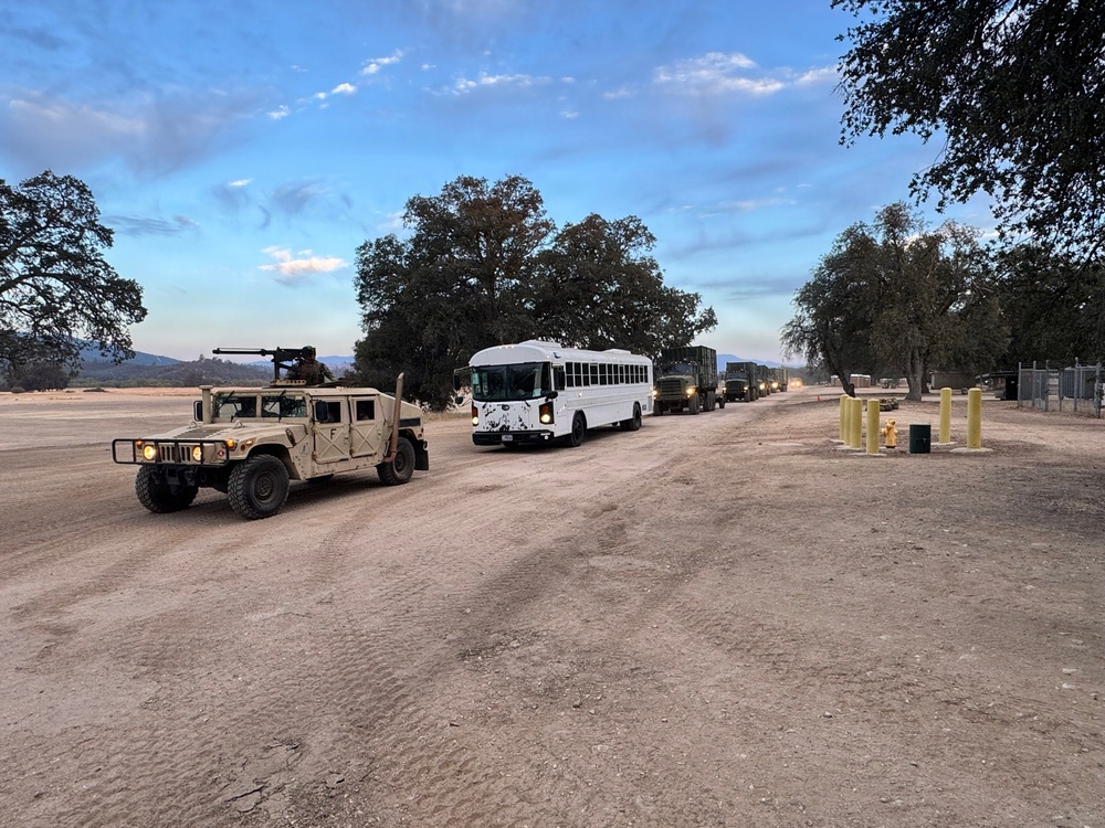 A military convoy is staged or moving along a dirt road at dusk or dawn, consisting of a lead HMMWV (Humvee) with a mounted weapon system, a white civilian-style bus, and multiple military trucks extending into the background. At least one soldier is visible manning the weapon mount on the Humvee. The setting appears to be a military installation or training area with sparse vegetation and open terrain.