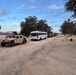 A military convoy is staged or moving along a dirt road at dusk or dawn, consisting of a lead HMMWV (Humvee) with a mounted weapon system, a white civilian-style bus, and multiple military trucks extending into the background. At least one soldier is visible manning the weapon mount on the Humvee. The setting appears to be a military installation or training area with sparse vegetation and open terrain.
