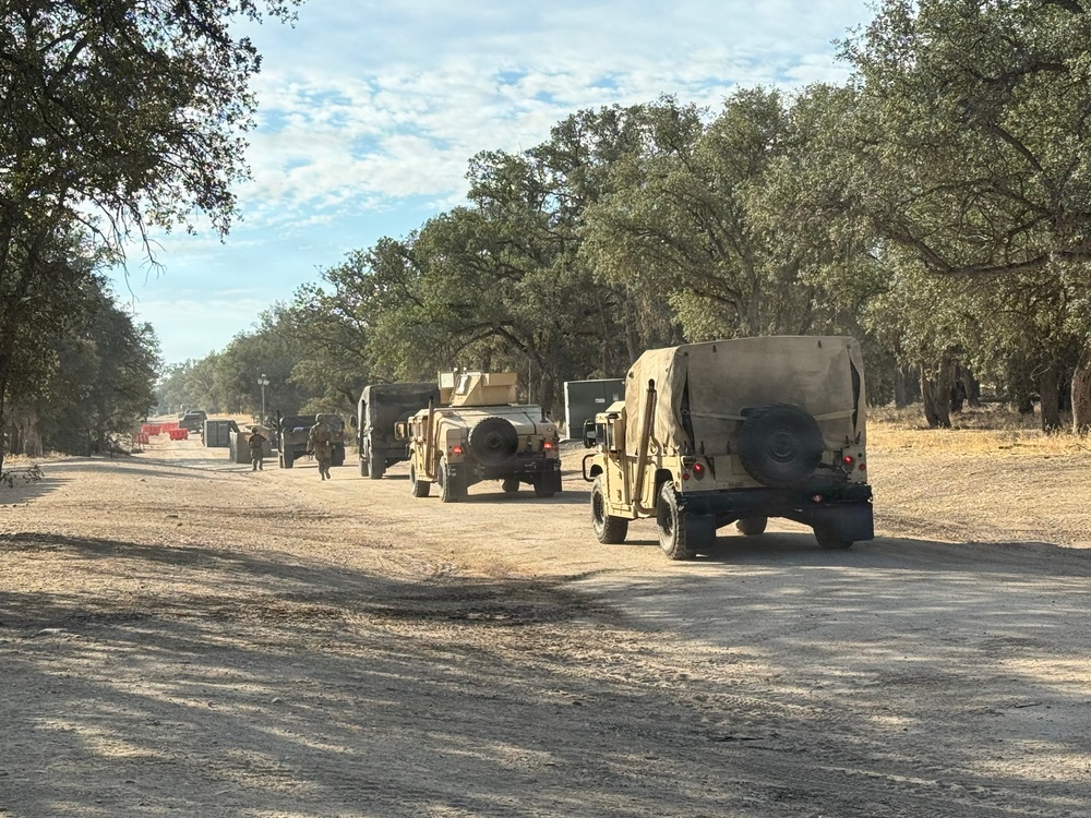 A military convoy of tan/desert-camouflage HMMWV (Humvee) vehicles and larger military trucks is staged or moving along a dry, unpaved dirt road lined with oak trees. The setting appears to be a training area or military installation in a dry, semi-arid environment, likely in California based on the oak tree landscape. At least one soldier on foot is visible near the vehicles.