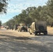 A military convoy of tan/desert-camouflage HMMWV (Humvee) vehicles and larger military trucks is staged or moving along a dry, unpaved dirt road lined with oak trees. The setting appears to be a training area or military installation in a dry, semi-arid environment, likely in California based on the oak tree landscape. At least one soldier on foot is visible near the vehicles.