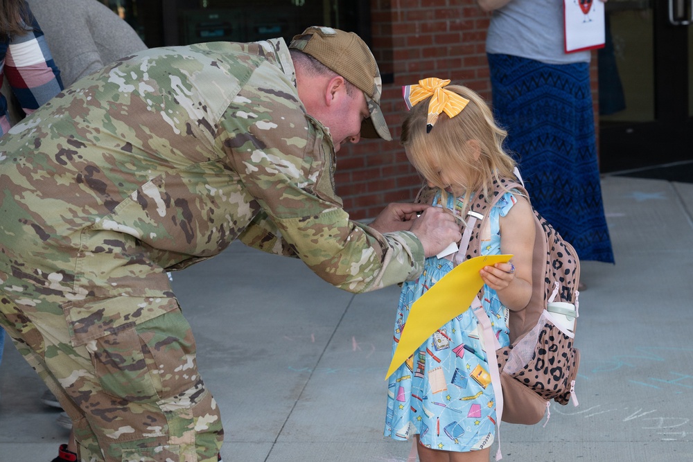 A military service member in full camouflage uniform bends down to interact with a young girl outside what appears to be a school entrance. The girl is wearing a light blue dress with school-supply prints, a yellow hair bow, and a leopard-print backpack, and is holding a yellow piece of paper. The scene suggests a school drop-off moment, possibly a surprise homecoming or first-day-of-school visit.