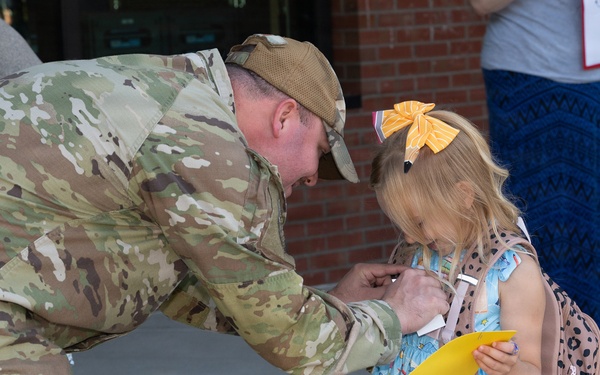 A military service member in full camouflage uniform bends down to interact with a young girl outside what appears to be a school entrance. The girl is wearing a light blue dress with school-supply prints, a yellow hair bow, and a leopard-print backpack, and is holding a yellow piece of paper. The scene suggests a school drop-off moment, possibly a surprise homecoming or first-day-of-school visit.