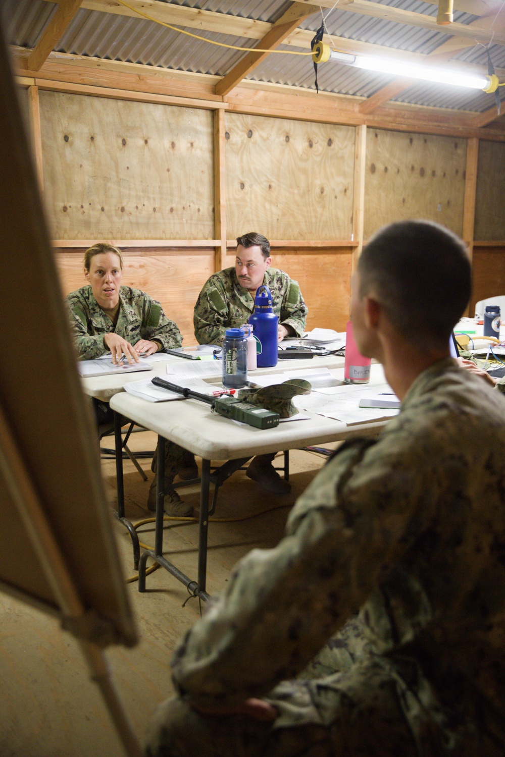 Three military personnel in camouflage uniforms are gathered around a folding table inside a temporary wooden structure, engaged in what appears to be a briefing or planning meeting. Two individuals seated at the table face a third person whose back is to the camera. The setting appears to be a forward operating base or field operations center.