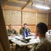 Three military personnel in camouflage uniforms are gathered around a folding table inside a temporary wooden structure, engaged in what appears to be a briefing or planning meeting. Two individuals seated at the table face a third person whose back is to the camera. The setting appears to be a forward operating base or field operations center.