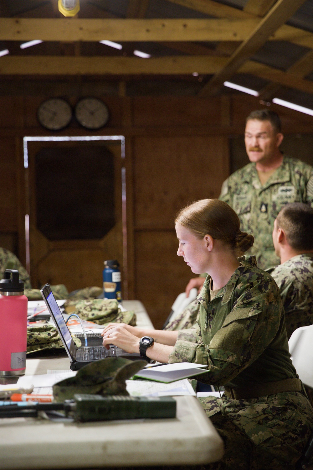 Three U.S. Navy personnel are working inside a wooden field operations building or temporary command post. A female sailor in the foreground is focused on a laptop computer, while two male sailors are visible in the background. The setting appears to be a forward operating or training environment based on the rustic wooden structure and field equipment present.