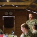 Three U.S. Navy personnel are working inside a wooden field operations building or temporary command post. A female sailor in the foreground is focused on a laptop computer, while two male sailors are visible in the background. The setting appears to be a forward operating or training environment based on the rustic wooden structure and field equipment present.