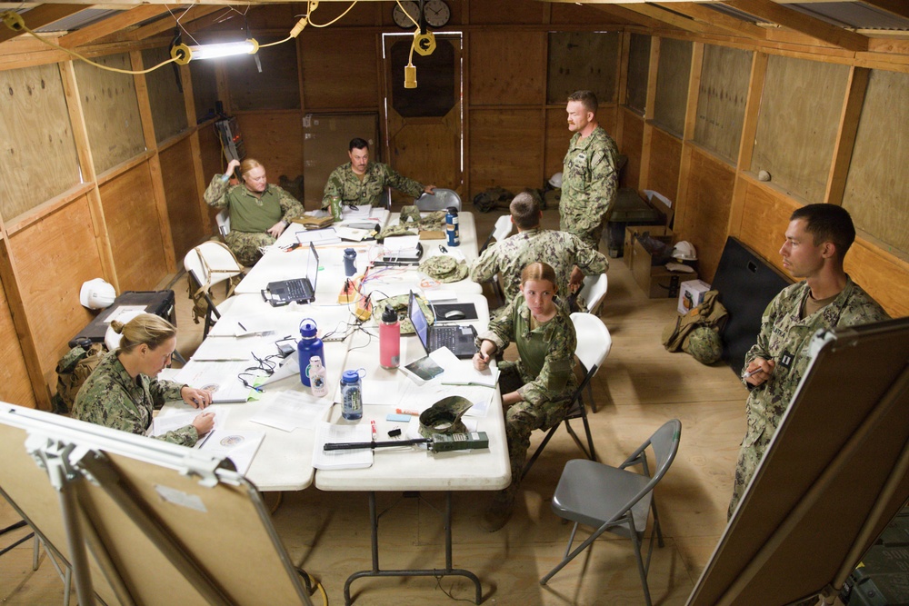 A group of approximately eight military personnel in camouflage uniforms are gathered in a temporary wooden structure that appears to be a field operations center or command post. They are seated and standing around folding tables covered with laptops, documents, water bottles, and various equipment. The setting suggests an active planning or briefing session in a forward operating environment.