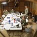 A group of approximately eight military personnel in camouflage uniforms are gathered in a temporary wooden structure that appears to be a field operations center or command post. They are seated and standing around folding tables covered with laptops, documents, water bottles, and various equipment. The setting suggests an active planning or briefing session in a forward operating environment.
