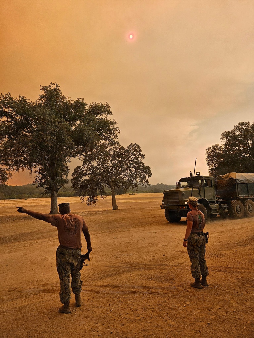 Two military personnel in camouflage uniforms are directing a large military cargo truck across a dusty open field under a heavily smoke-filled orange sky. The scene strongly suggests wildfire response operations, with the sun barely visible as a dim red orb through thick smoke haze. The environment shows dry, arid terrain with scattered oak trees typical of California's inland regions.