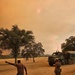 Two military personnel in camouflage uniforms are directing a large military cargo truck across a dusty open field under a heavily smoke-filled orange sky. The scene strongly suggests wildfire response operations, with the sun barely visible as a dim red orb through thick smoke haze. The environment shows dry, arid terrain with scattered oak trees typical of California's inland regions.