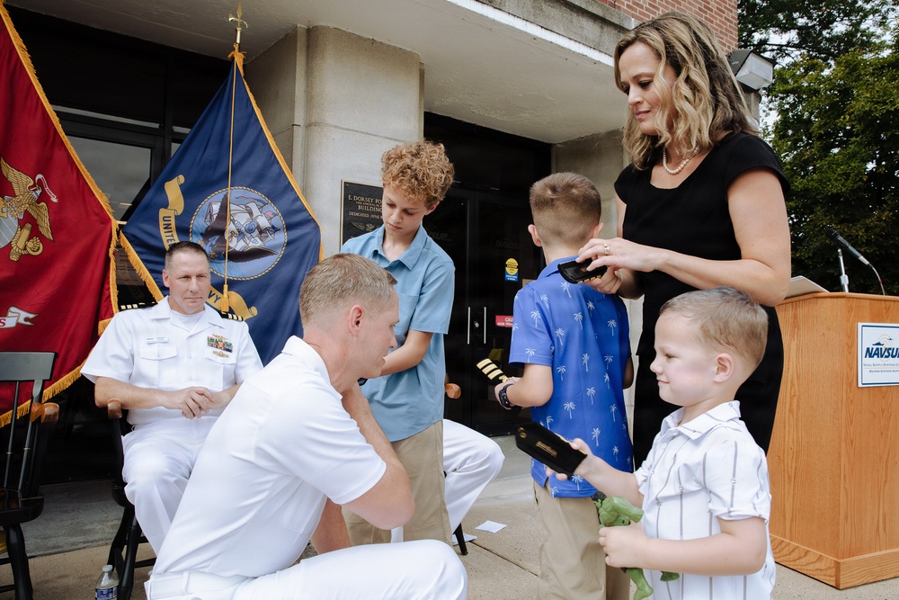 A military promotion or retirement ceremony is taking place outdoors in front of a building, likely a Naval facility based on the NAVSUP podium visible in the scene. A Navy officer in white dress uniform kneels down to interact with his children while his wife assists with what appears to be pinning new rank insignia. A senior officer in white uniform observes from a seated position in the background.