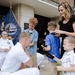 A military promotion or retirement ceremony is taking place outdoors in front of a building, likely a Naval facility based on the NAVSUP podium visible in the scene. A Navy officer in white dress uniform kneels down to interact with his children while his wife assists with what appears to be pinning new rank insignia. A senior officer in white uniform observes from a seated position in the background.