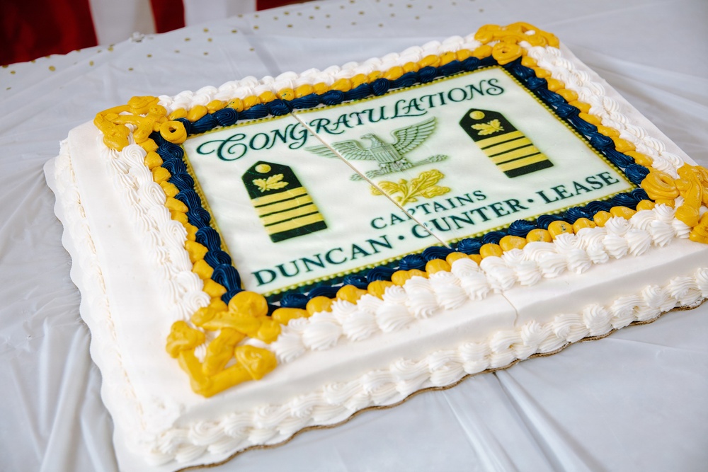 A large rectangular sheet cake celebrating a military promotion ceremony for three individuals being promoted to the rank of Captain. The cake features Navy Captain insignia including eagle devices, rank stripes, and anchor decorations in navy blue and gold frosting. The names Duncan, Gunter, and Lease are displayed prominently on the cake's fondant topper.