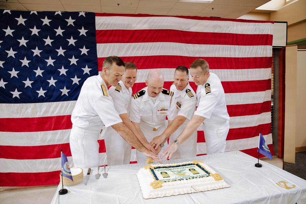 Five U.S. Navy officers in white dress uniforms are gathered around a table cutting a congratulatory cake in front of a large American flag backdrop. The ceremony appears to be a military retirement or promotion celebration, as indicated by the 'Congratulations' text visible on the cake. The setting appears to be an indoor military facility decorated for the formal occasion.