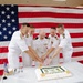 Five U.S. Navy officers in white dress uniforms are gathered around a table cutting a congratulatory cake in front of a large American flag backdrop. The ceremony appears to be a military retirement or promotion celebration, as indicated by the 'Congratulations' text visible on the cake. The setting appears to be an indoor military facility decorated for the formal occasion.