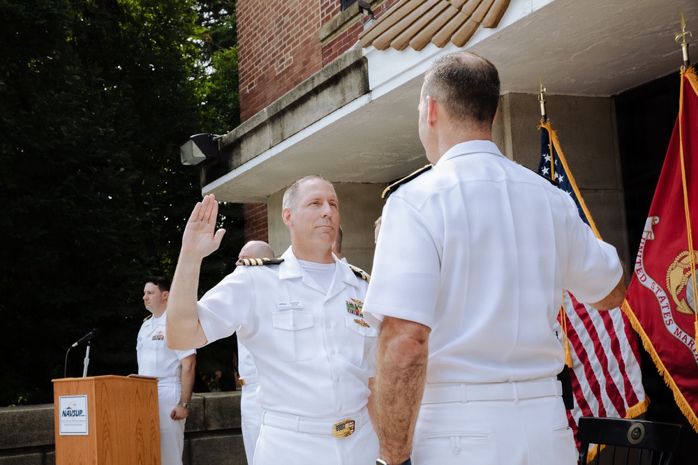 A U.S. Navy military ceremony is taking place outdoors, likely a promotion or oath of office ceremony. A naval officer in white dress uniform is raising his right hand, apparently taking an oath administered by a senior officer facing away from the camera. The United States flag and United States Marine Corps flag are visible in the background alongside a NAVSUP podium.