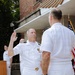A U.S. Navy military ceremony is taking place outdoors, likely a promotion or oath of office ceremony. A naval officer in white dress uniform is raising his right hand, apparently taking an oath administered by a senior officer facing away from the camera. The United States flag and United States Marine Corps flag are visible in the background alongside a NAVSUP podium.