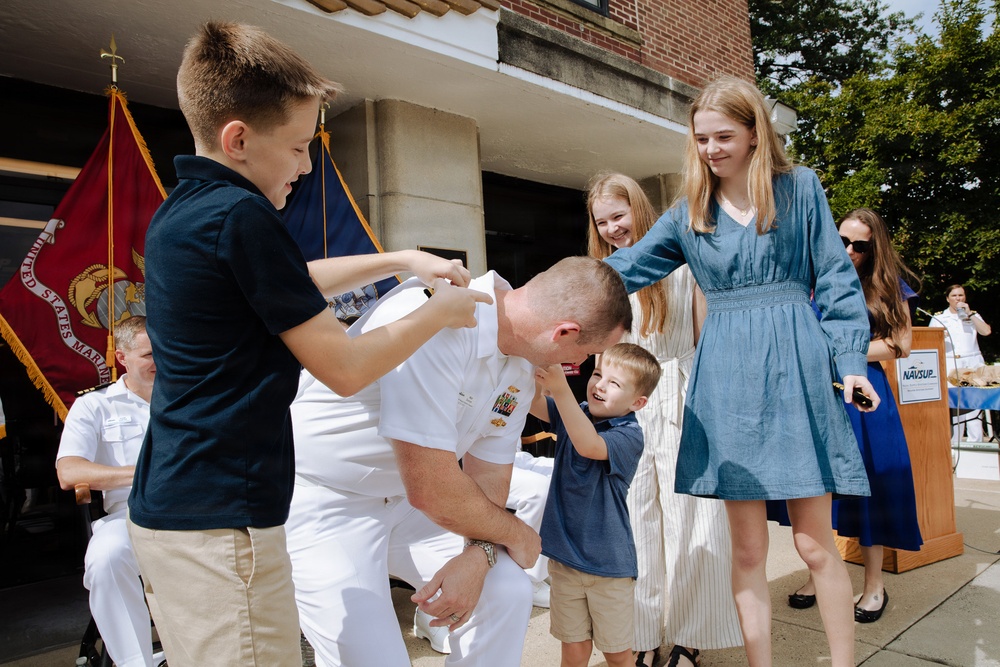 A U.S. Navy officer in white dress uniform is being embraced and pinned by his children during what appears to be a military promotion or retirement ceremony. The scene takes place outdoors in front of a brick building, with a United States Marine Corps flag visible in the background. The moment is joyful and celebratory, with multiple children participating in the pinning tradition.